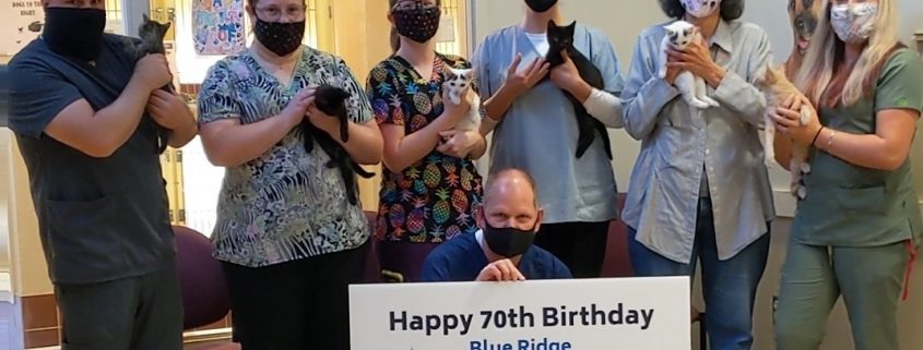 a group of men and women in scrubs stand holding cats, while wearing face masks. A man kneels in front holding a sign with a cat and dog on it saying "happy 70th birthday Blue Ridge Humane Society"