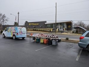 image of a brightly decorated van with tables set up behind it with piles of bagged pet food. In the background in a modern looking one story building with "boys and girls club of Henderson County" written on it.