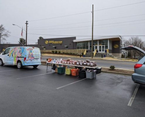 image of a brightly decorated van with tables set up behind it with piles of bagged pet food. In the background in a modern looking one story building with "boys and girls club of Henderson County" written on it.