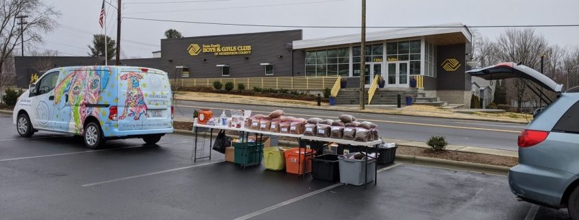 image of a brightly decorated van with tables set up behind it with piles of bagged pet food. In the background in a modern looking one story building with "boys and girls club of Henderson County" written on it.