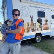 A man in an orange vest holds a small scruffy dog in front of a BRHS van