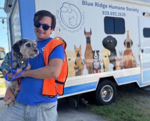 A man in an orange vest holds a small scruffy dog in front of a BRHS van