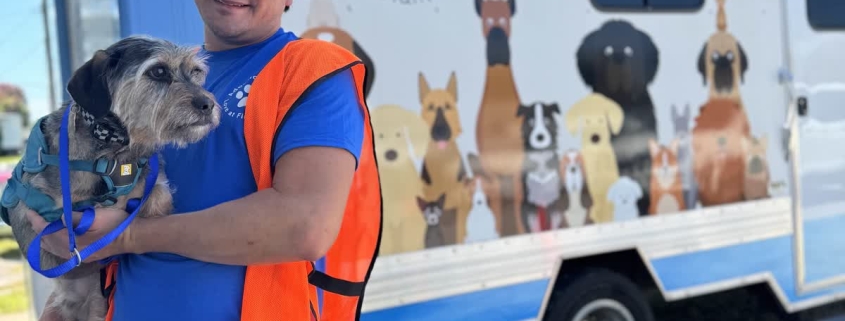 A man in an orange vest holds a small scruffy dog in front of a BRHS van